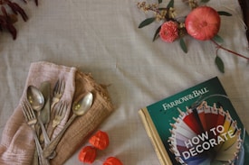 A collection of silverware including forks and spoons rests on a beige cloth on the left side, alongside a rustic book titled 'Farrow & Ball: How to Decorate' on the right. Above the book, there are decorative elements, including a pink flower and a pomegranate, accompanied by some green foliage. The setting gives a vintage and cozy feel.