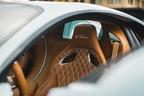 The interior of a luxury car featuring a brown leather sport seat with diamond-patterned stitching and the word 'Chiron' embroidered on the headrest. The car's interior displays sleek curves and metallic accents, emphasizing a sense of refinement and exclusivity.