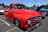 A vibrant red vintage truck is parked in an outdoor setting, reflecting sunlight on its glossy surface. The truck has a classic, retro design with rounded headlights and chrome details. Surrounding the truck are other vintage vehicles, including a blue car with an open hood. The scene conveys a car show or vintage vehicle gathering under clear blue skies.