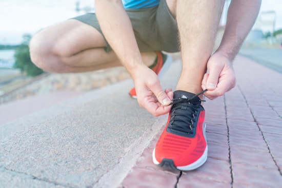 A person is crouching down, tying the laces of a red athletic shoe on a paved surface. The background includes an out-of-focus outdoor setting.