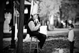 An older man is sitting on a stool playing a wind instrument, while a young child stands nearby, observing him. The scene is set outdoors on a street covered in fallen leaves, adding an autumn atmosphere. Both individuals are dressed warmly, suggesting cold weather.