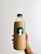 A hand holds a plastic bottle filled with a light brown liquid, featuring a black cap. The bottle has a green circular label with a white silhouette of a person's profile.