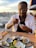 A person with glasses and a white shirt is enjoying a meal of oysters at an outdoor setting, possibly by a harbor or marina. The table is set with a glass, cutlery, and a plate of oyster shells, garnished with greens and a lemon wedge. The background is softly blurred, suggesting a warm, serene environment.