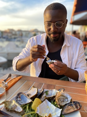 A person with glasses and a white shirt is enjoying a meal of oysters at an outdoor setting, possibly by a harbor or marina. The table is set with a glass, cutlery, and a plate of oyster shells, garnished with greens and a lemon wedge. The background is softly blurred, suggesting a warm, serene environment.