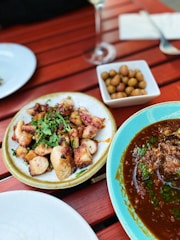 A wooden table set with a variety of dishes, including a plate of grilled octopus garnished with herbs, a dish of olives, and a bowl of rich, spicy stew garnished with green herbs. The scene suggests an outdoor dining experience.