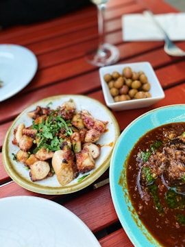 A wooden table set with a variety of dishes, including a plate of grilled octopus garnished with herbs, a dish of olives, and a bowl of rich, spicy stew garnished with green herbs. The scene suggests an outdoor dining experience.