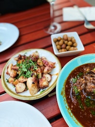A wooden table set with a variety of dishes, including a plate of grilled octopus garnished with herbs, a dish of olives, and a bowl of rich, spicy stew garnished with green herbs. The scene suggests an outdoor dining experience.