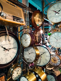A collection of vintage wall clocks and an old-fashioned radio are featured, with various sizes and styles of clocks in a luxurious metallic finish. The clocks display Roman numerals, and the surrounding environment indicates a shop or market setting.