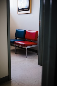 A partially open door reveals a small waiting area with a pair of chairs, one with dark blue upholstery and the other with red. Above the chairs, a framed picture hangs on the wall. The floor is covered with light-colored linoleum tiles.