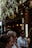 A group of people stand outside a pizzeria, with a sign above the door that reads 'Pizzeria'. The entrance is partially covered by flowering plants hanging overhead. The scene is bustling with activity as several individuals wearing sunglasses and casual clothing are visible in the foreground. Warm lighting and decorative lamps create a cozy atmosphere inside the venue.