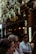 A group of people stand outside a pizzeria, with a sign above the door that reads 'Pizzeria'. The entrance is partially covered by flowering plants hanging overhead. The scene is bustling with activity as several individuals wearing sunglasses and casual clothing are visible in the foreground. Warm lighting and decorative lamps create a cozy atmosphere inside the venue.