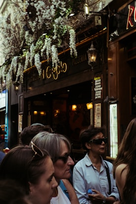A group of people stand outside a pizzeria, with a sign above the door that reads 'Pizzeria'. The entrance is partially covered by flowering plants hanging overhead. The scene is bustling with activity as several individuals wearing sunglasses and casual clothing are visible in the foreground. Warm lighting and decorative lamps create a cozy atmosphere inside the venue.