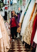 A person standing in a fabric store surrounded by various colorful and patterned textiles. The person is dressed in casual attire, raising a hand towards the camera as if gesturing or blocking the view.