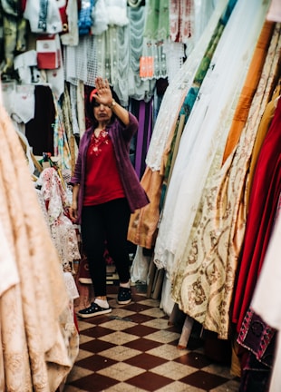 A person standing in a fabric store surrounded by various colorful and patterned textiles. The person is dressed in casual attire, raising a hand towards the camera as if gesturing or blocking the view.