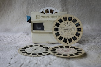 A vintage View-Master stereoscope viewer is shown alongside several view reels. The viewer is off-white with a blue lever, and the reels are labeled with destinations or themes, one mentioning Disneyland. The items are placed on a textured, neutral-colored surface, giving a nostalgic feel.