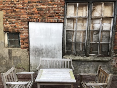 Outdoor patio setting with aged wooden furniture, including a table and two chairs, set in front of a rustic brick wall. The brick wall features two windows with distressed, cloudy glass panes and a weathered appearance.