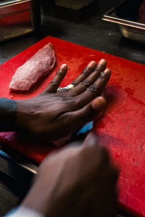 A piece of raw meat lies on a red cutting board, with a hand positioned next to it as if guiding a knife. The environment appears to be a kitchen setting with metal containers visible in the background.