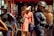 A group of police officers wearing protective gear, including helmets and body armor, stand near a woman holding a bag and shielding her eyes from the sun. Another person in a red tank top is visible nearby, holding a water bottle. The setting appears to be a busy street with a restaurant or shop in the background.