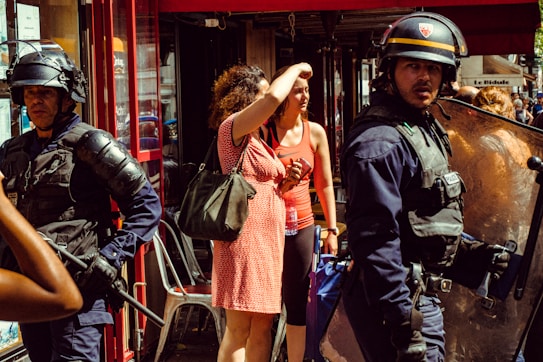 A group of police officers wearing protective gear, including helmets and body armor, stand near a woman holding a bag and shielding her eyes from the sun. Another person in a red tank top is visible nearby, holding a water bottle. The setting appears to be a busy street with a restaurant or shop in the background.