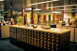 A library with a vintage card catalog system in the foreground, surrounded by bookshelves and modern furniture. Several potted plants are placed atop the counter and around the room, adding a touch of greenery. The lighting is warm, and the overall atmosphere is quiet and studious, with neatly organized books and files lining the shelves in the background.