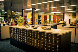 A library with a vintage card catalog system in the foreground, surrounded by bookshelves and modern furniture. Several potted plants are placed atop the counter and around the room, adding a touch of greenery. The lighting is warm, and the overall atmosphere is quiet and studious, with neatly organized books and files lining the shelves in the background.