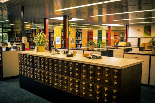 A library with a vintage card catalog system in the foreground, surrounded by bookshelves and modern furniture. Several potted plants are placed atop the counter and around the room, adding a touch of greenery. The lighting is warm, and the overall atmosphere is quiet and studious, with neatly organized books and files lining the shelves in the background.