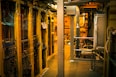 A dimly lit server room with multiple racks of computer servers surrounded by cables and various technical equipment. The space has an industrial feel with visible pipes and ductwork, and a sign on one rack stating 'Do not use'.