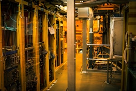 A dimly lit server room with multiple racks of computer servers surrounded by cables and various technical equipment. The space has an industrial feel with visible pipes and ductwork, and a sign on one rack stating 'Do not use'.