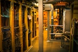 A dimly lit server room with multiple racks of computer servers surrounded by cables and various technical equipment. The space has an industrial feel with visible pipes and ductwork, and a sign on one rack stating 'Do not use'.