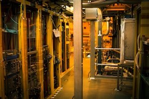 A dimly lit server room with multiple racks of computer servers surrounded by cables and various technical equipment. The space has an industrial feel with visible pipes and ductwork, and a sign on one rack stating 'Do not use'.