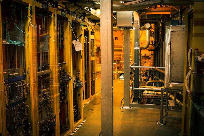 A dimly lit server room with multiple racks of computer servers surrounded by cables and various technical equipment. The space has an industrial feel with visible pipes and ductwork, and a sign on one rack stating 'Do not use'.