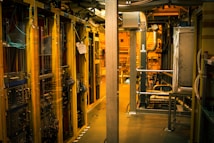 A dimly lit server room with multiple racks of computer servers surrounded by cables and various technical equipment. The space has an industrial feel with visible pipes and ductwork, and a sign on one rack stating 'Do not use'.