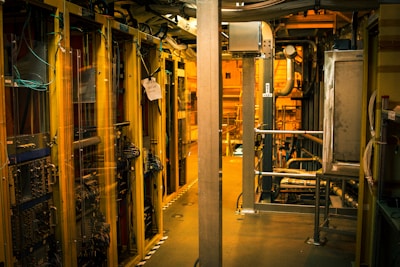 A dimly lit server room with multiple racks of computer servers surrounded by cables and various technical equipment. The space has an industrial feel with visible pipes and ductwork, and a sign on one rack stating 'Do not use'.