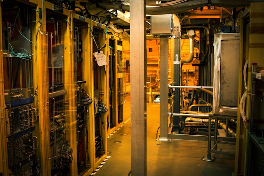 A dimly lit server room with multiple racks of computer servers surrounded by cables and various technical equipment. The space has an industrial feel with visible pipes and ductwork, and a sign on one rack stating 'Do not use'.