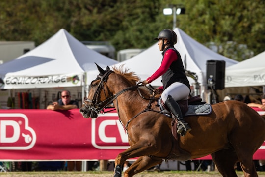 A person riding a brown horse during an equestrian event, wearing a helmet and riding gear. White tents and spectators are visible in the background, indicating a public setting.