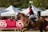 A person riding a brown horse during an equestrian event, wearing a helmet and riding gear. White tents and spectators are visible in the background, indicating a public setting.