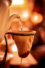 A warm-toned image captures the process of pouring hot water from a kettle over a cloth filter. The scene is illuminated by a soft, ambient glow, creating a cozy atmosphere as the liquid slowly drips through the filter.