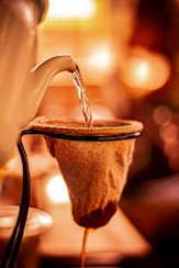 A warm-toned image captures the process of pouring hot water from a kettle over a cloth filter. The scene is illuminated by a soft, ambient glow, creating a cozy atmosphere as the liquid slowly drips through the filter.