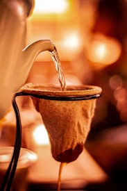 A warm-toned image captures the process of pouring hot water from a kettle over a cloth filter. The scene is illuminated by a soft, ambient glow, creating a cozy atmosphere as the liquid slowly drips through the filter.