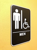 A restroom sign indicating facilities for men and accessibility for individuals with disabilities. The sign features simple icons of a person and a wheelchair user, and is mounted on a yellow wall.