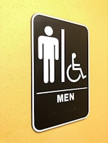 A restroom sign indicating facilities for men and accessibility for individuals with disabilities. The sign features simple icons of a person and a wheelchair user, and is mounted on a yellow wall.