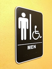 A restroom sign indicating facilities for men and accessibility for individuals with disabilities. The sign features simple icons of a person and a wheelchair user, and is mounted on a yellow wall.