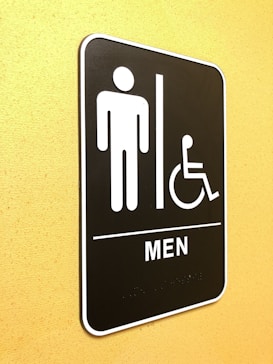 A restroom sign indicating facilities for men and accessibility for individuals with disabilities. The sign features simple icons of a person and a wheelchair user, and is mounted on a yellow wall.