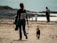 A person with a patterned backpack walks alongside a small brown dog on a sandy beach. The person carries a blue dog toy and wears casual clothing. In the distance, there are blurred figures of other people along the shoreline and rocky areas.