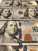 A collection of stacked United States 100 dollar bills, featuring a portrait of Benjamin Franklin. The bills display various intricate designs, watermarks, and security features like blue security ribbons and holograms.