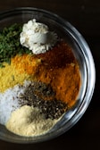 A clear glass bowl is filled with various types of ground spices and herbs. The ingredients inside the bowl include green leafy herbs, a mound of white flour, yellow turmeric, red chili powder, black pepper, and coarse salt. Each component retains its distinct color and texture, creating a visually appealing contrast of colors.