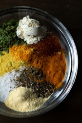A clear glass bowl is filled with various types of ground spices and herbs. The ingredients inside the bowl include green leafy herbs, a mound of white flour, yellow turmeric, red chili powder, black pepper, and coarse salt. Each component retains its distinct color and texture, creating a visually appealing contrast of colors.