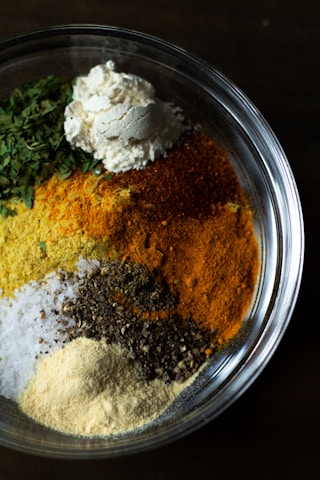 A clear glass bowl is filled with various types of ground spices and herbs. The ingredients inside the bowl include green leafy herbs, a mound of white flour, yellow turmeric, red chili powder, black pepper, and coarse salt. Each component retains its distinct color and texture, creating a visually appealing contrast of colors.