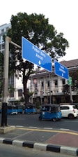 Several blue autorickshaws are parked along a street with large blue road signs pointing toward Pasar Baru and Ancol. The background features trees and a multi-story building.