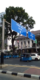 Several blue autorickshaws are parked along a street with large blue road signs pointing toward Pasar Baru and Ancol. The background features trees and a multi-story building.